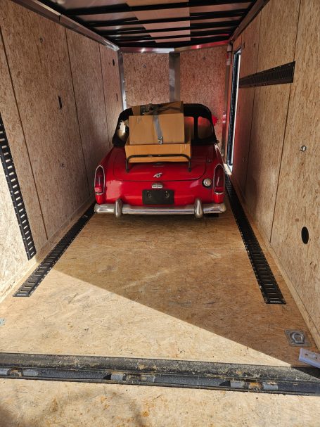 Red car parked inside a trailer with a box on the back seat.