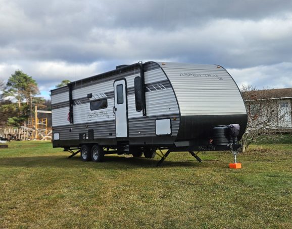 A modern travel trailer parked on green grass with trees and a building in the background.